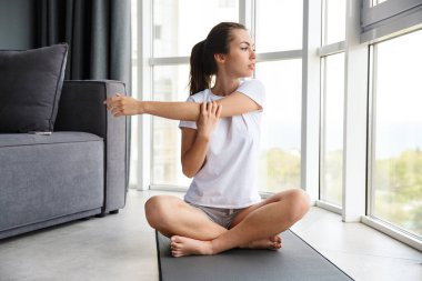 Pencerenin yanındaki yoga minderinde otururken kolunu uzatan odaklanmış genç bir kadın görüntüsü