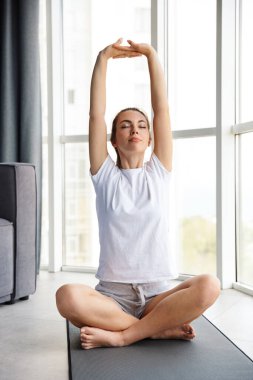 Pencerenin yanındaki yoga minderinde otururken egzersiz yapan odaklanmış genç bir kadın görüntüsü