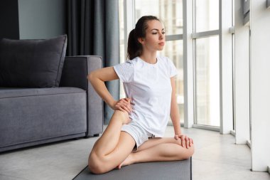 Pencerenin yanındaki yoga minderinde otururken egzersiz yapan odaklanmış genç bir kadın görüntüsü