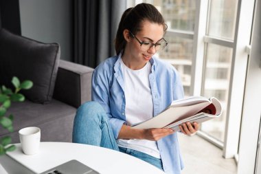 Gülümseyen, kitap okuyan ve masada otururken dizüstü bilgisayarla çalışan güzel bir kadın resmi.