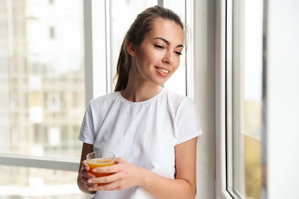 窓の近くに立ちながら笑顔で新鮮なジュースを飲んでいる幸せな若い素敵な女性のイメージ屋内 — ストック写真