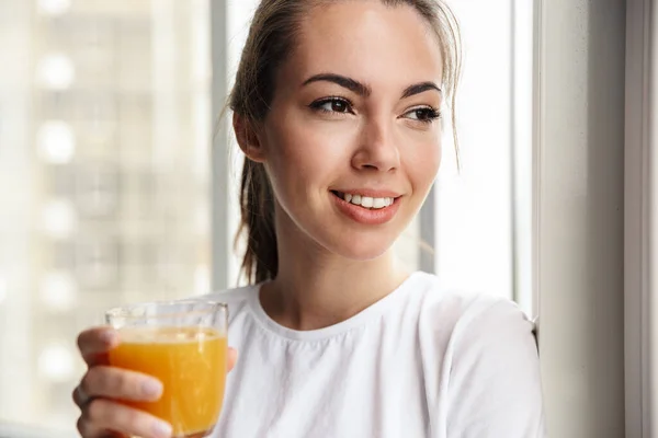 窓の近くに立ちながら笑顔で新鮮なジュースを飲んでいる幸せな若い素敵な女性のイメージ屋内 — ストック写真