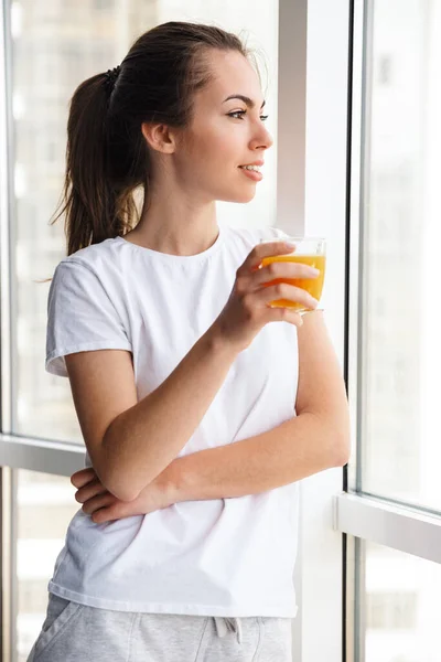 站在室内靠窗的时候 高兴而和善的年轻女子笑着喝新鲜果汁的照片 — 图库照片