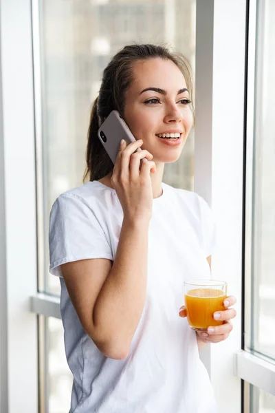 图片中 一个微笑的女人站在室内 一边用手机一边喝果汁一边说话 — 图库照片