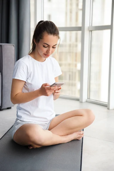 Obrázek Usmívající Mladé Ženy Pomocí Mobilu Při Sezení Podložce Jógy — Stock fotografie