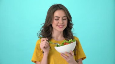 Sarı tişört giyen gülümseyen genç bir kadın yeşil salata yiyor. Mavi arka planda tek başına duruyor.