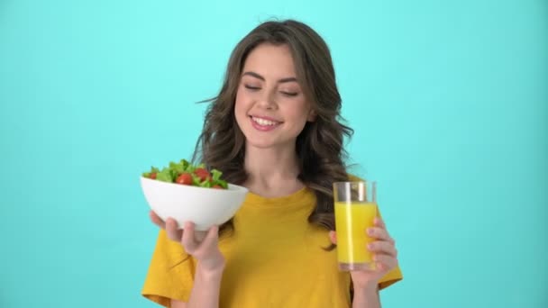 一位穿着黄色T恤的可爱的年轻女子 手里拿着一碗沙拉和新鲜果汁 与蓝色的背景隔离开来 — 图库视频影像