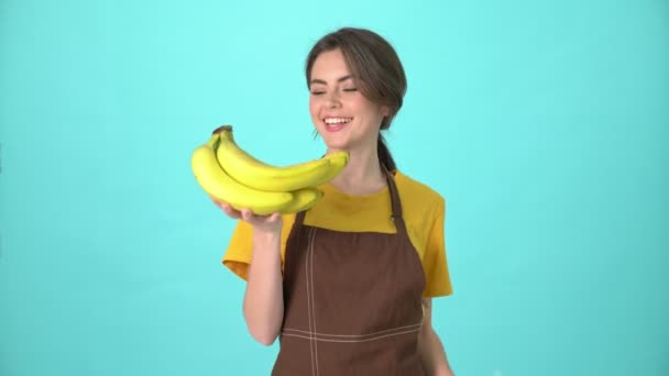 茶色のエプロンをした幸せな若い女性が青い背景の上に隔離されたバナナスタンドを保持しています — ストック動画