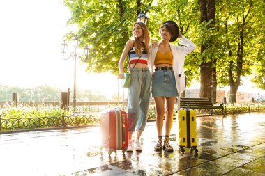 Yeşil şehir bulvarında bavullarla yürürken gülümseyen ve sarılan iki güzel kadının fotoğrafı.