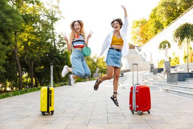 İki çekici turist kadının parkta bavullarla yürürken gülüp eğlendikleri bir fotoğraf.