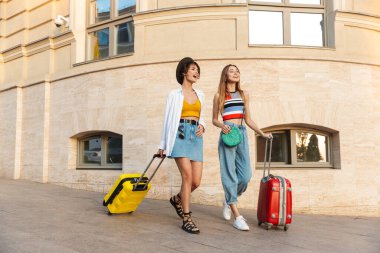 İki güzel turist kadının fotoğrafı, gülümseyen ve valizlerle şehir caddesinde yürürken.