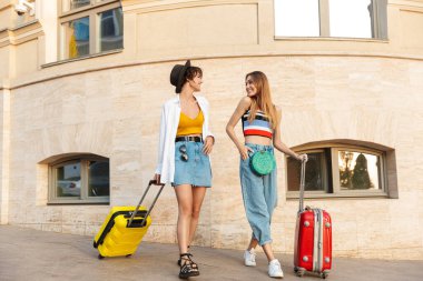 İki Kafkasyalı turist kadının fotoğrafı. Şehrin caddesinde bavullarıyla gülümsüyor ve yürüyorlar.
