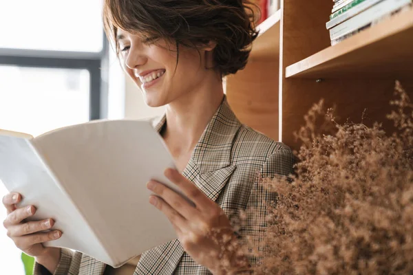 Smiling Young Woman Entrepreneur Reading Book While Standing Office Bookshelf — Stock Photo, Image