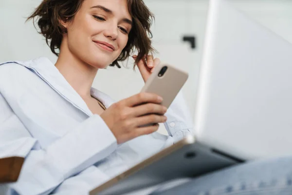 若い魅力的な女性起業家の笑顔を自信を持ってオフィスの椅子に座って ラップトップコンピュータで作業し 携帯電話を使用して — ストック写真