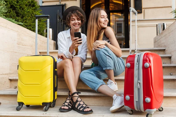 ホテルの近くのスーツケースと階段に座っている間 携帯電話を使用して2人の笑顔の白人女性の写真 — ストック写真