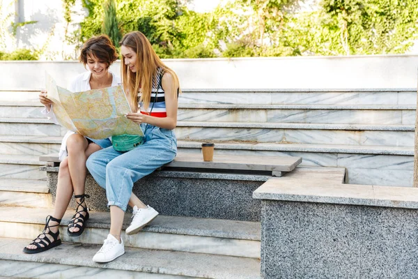 两个微笑的年轻女子坐在靠近城市街道楼梯的长椅上 手握地图 看着地图的照片 — 图库照片