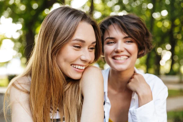 緑の公園のベンチに腰を下ろして笑っている若い女性2人の写真 — ストック写真
