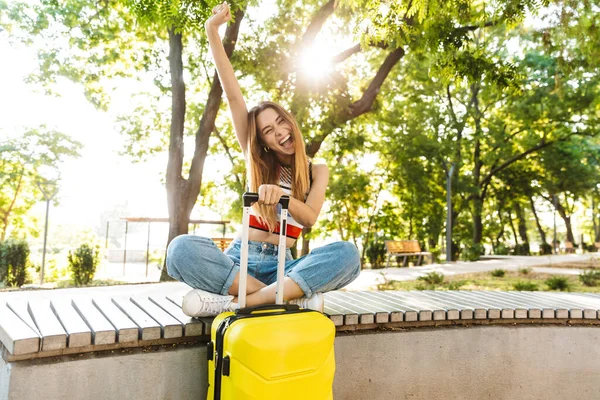 照片中 一个快乐的旅游女孩坐在绿地公园的长椅上 一边笑一边提行李 — 图库照片