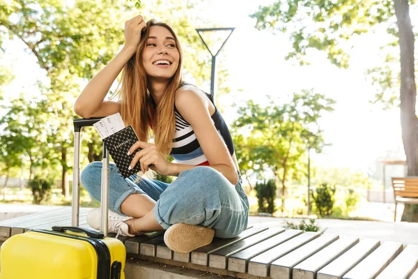 照片中 一个快乐的旅游女孩坐在绿园的长椅上 背着行李 手里拿着护照和旅游票 — 图库照片