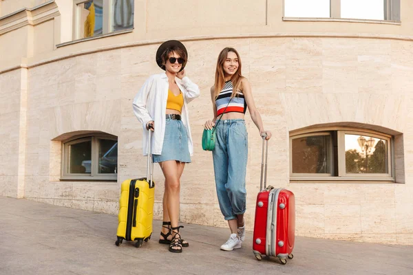 照片中 两个漂亮的旅游女孩带着手提箱在城市街道上散步时微笑着 — 图库照片