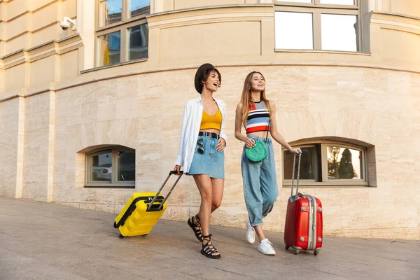 两个漂亮的女游客笑着带着手提箱走在城市街道上的照片 — 图库照片