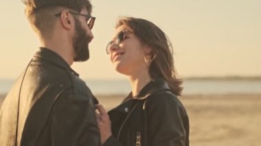 Gün batımında sahilde yürürken gülümseyen genç bir çiftin birbirlerine sarılıp baktıkları yakın çekim görüntüsü.