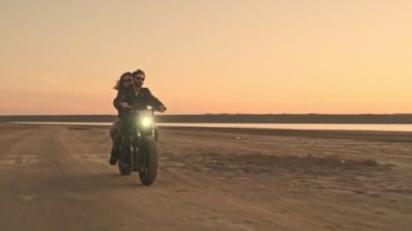 Güneş gözlüğü takan genç bir çift gün batımında plajda birlikte motosiklet sürüyorlar.