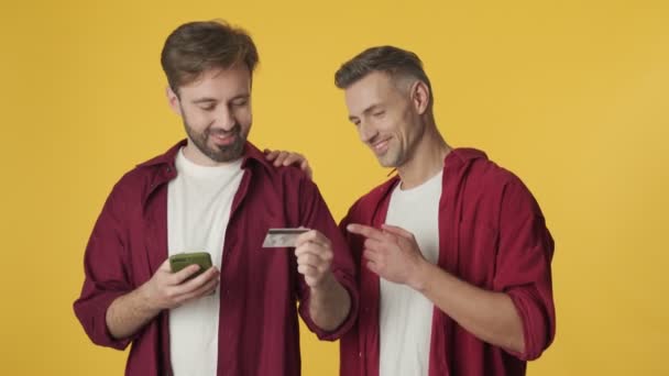 英俊的成年朋友们一边用智能手机和信用卡购物 一边站在黄色的背景下孤立无援 — 图库视频影像