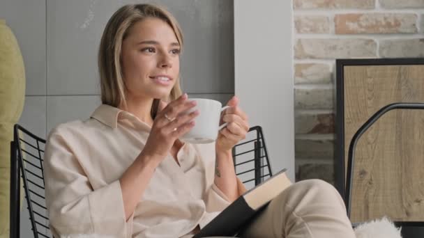 魅力的なブロンド女性飲むお茶と見ながら離れて座っています本の上の椅子に家で — ストック動画