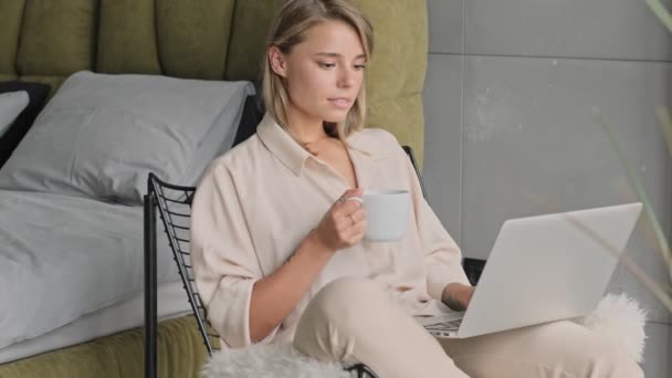 满意的金发女人坐在椅子上喝茶时使用笔记本电脑 — 图库视频影像