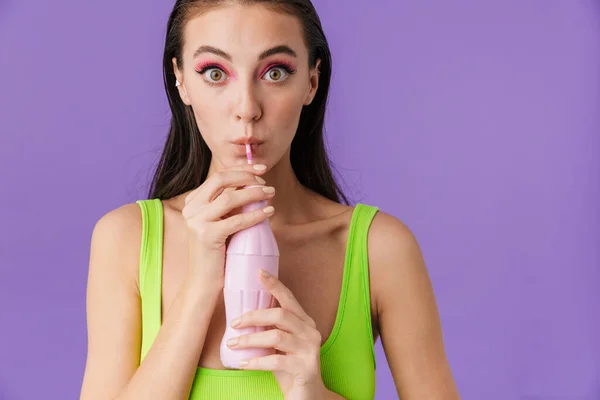 若い驚きの女性がペットボトルからソーダを飲み 紫色の背景に隔離されたカメラを見る写真 — ストック写真
