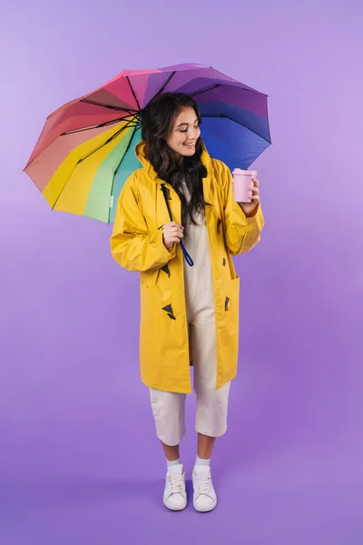 Image of positive brunette woman in yellow raincoat posing isolated over purple wall background holding umbrella and coffee.