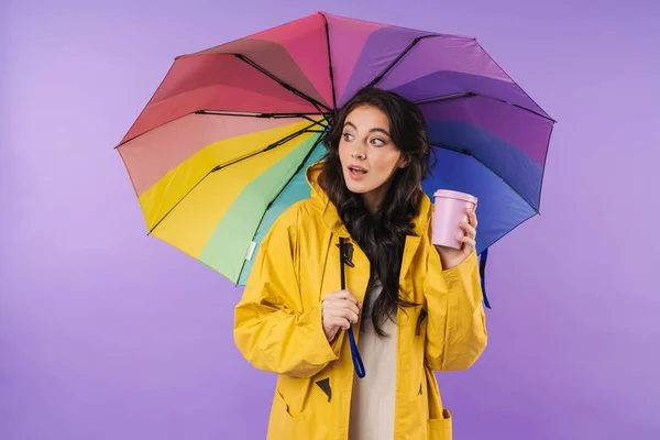 Image of concentrated brunette woman in yellow raincoat posing isolated over purple wall background holding umbrella and coffee looking aside.