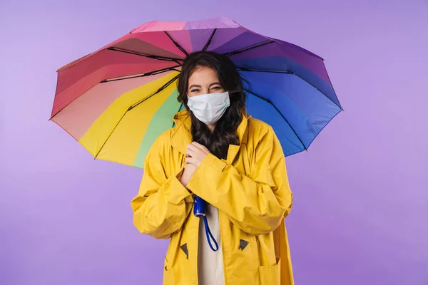 Image of positive happy woman in yellow raincoat posing isolated over purple wall background holding umbrella wearing medical mask.