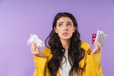 Mor duvar arkasına izole edilmiş, elinde peçete ve haplarla üzgün ve hasta bir kadın görüntüsü..