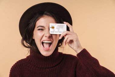 Mutlu, duygusal, esmer bir kadının bej duvar arkasında kredi kartı tutarak poz vermesi..
