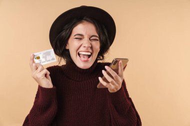 Çığlık atan, duygusal, esmer bir kadının bej duvar arkasında poz verirken görüntüsü. Elinde cep telefonu ile kredi kartı tutarken..