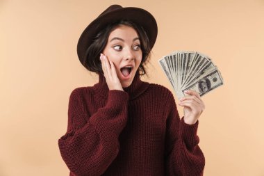 Image of young surprised emotional brunette woman isolated over beige wall background holding money. clipart