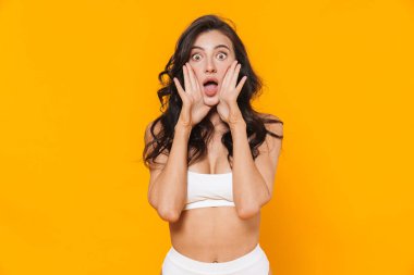 Sarı duvarın üzerinden izole edilmiş beyaz mayo giymiş, kameraya poz veren ve bağıran heyecanlı genç bir kadın resmi.