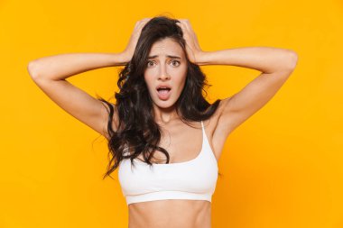 Mayo giymiş, başını tutup sarı duvarın üzerinden izole edilmiş kameraya bakan genç bir kadının görüntüsü.