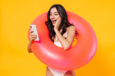 Heyecanlı, lastik halkalı, cep telefonu kullanan ve sarı duvarda izole edilmiş gülümseyen bir kadın resmi.