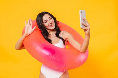Sarı duvarda izole edilmiş cep telefonundan selfie çeken lastik halkalı neşeli bir kadın resmi.