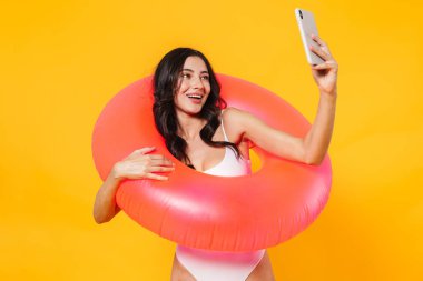 Sarı duvardan izole edilmiş cep telefonunda gülümseyen ve selfie çeken kauçuk yüzüklü neşeli bir kadın resmi.