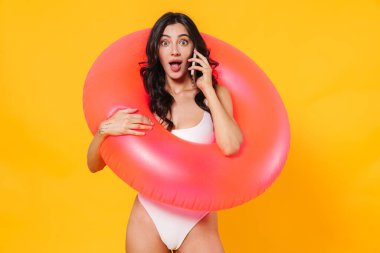 Sarı duvardan izole edilmiş cep telefonuyla konuşan kauçuk yüzüklü, şaşırmış genç bir kadın.