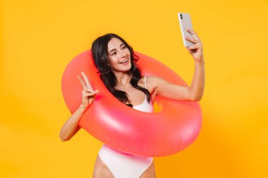 Lastik halkalı, barış işareti yapan ve sarı duvarda izole edilmiş cep telefonundan selfie çeken neşeli bir kadın resmi.