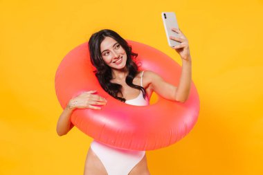 Sarı duvardan izole edilmiş cep telefonunda gülümseyen ve selfie çeken kauçuk yüzüklü neşeli bir kadın resmi.