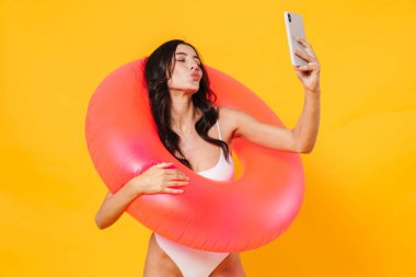 Lastik yüzüklü, öpücük dudaklı ve sarı duvarda izole edilmiş cep telefonundan selfie çeken neşeli bir kadın resmi.