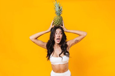 Sarı duvarın üzerinde ananas tutan mayolu, şaşırmış esmer kadın resmi.