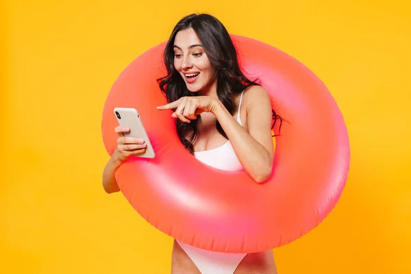 Image of cheerful woman with rubber ring using and pointing finger at cellphone isolated over yellow wall