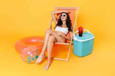 Sarı duvarda izole edilmiş şezlongda dinlenirken mayo giymiş mutlu bir kadının görüntüsü.